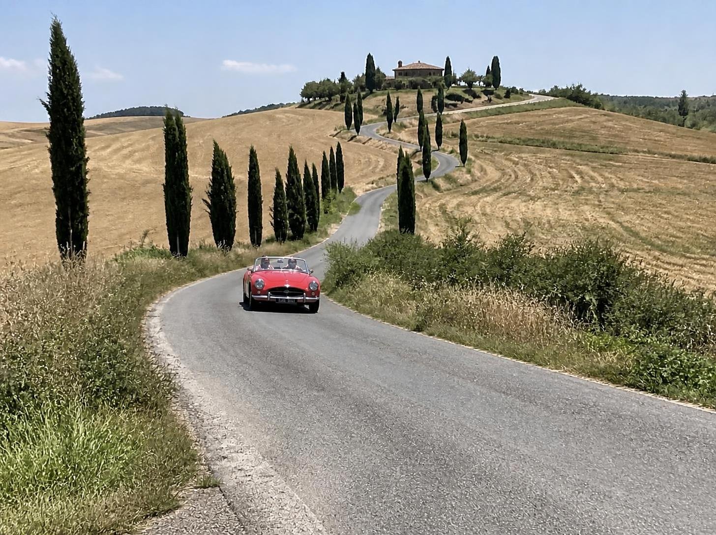 A red classic convertible on a winding road through Val d'Orcia